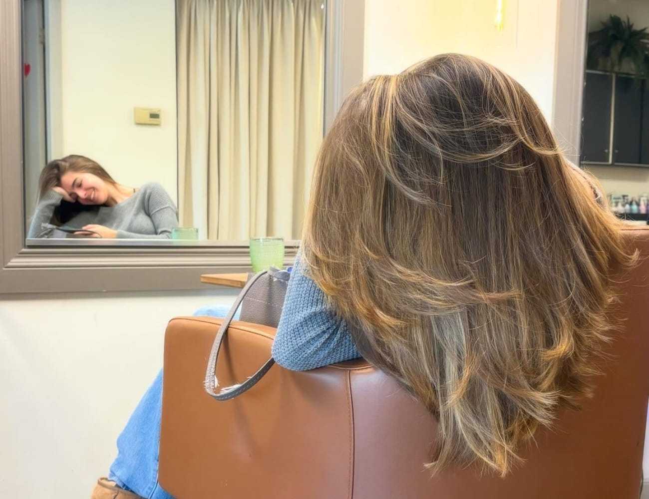 Woman with long hair sitting in a salon chair, smiling into a mirror.