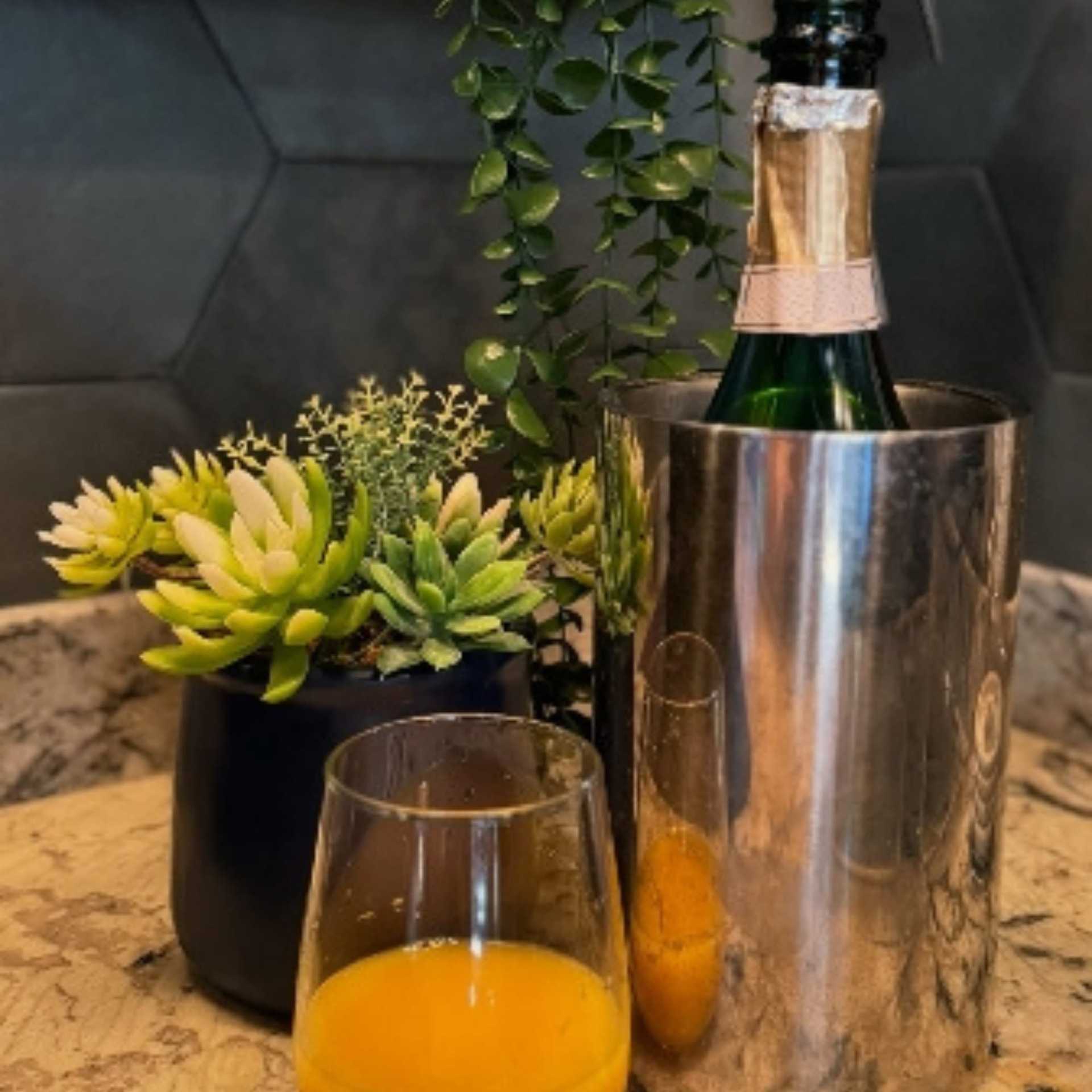 Champagne in an ice bucket next to a glass of orange juice and a small plant.