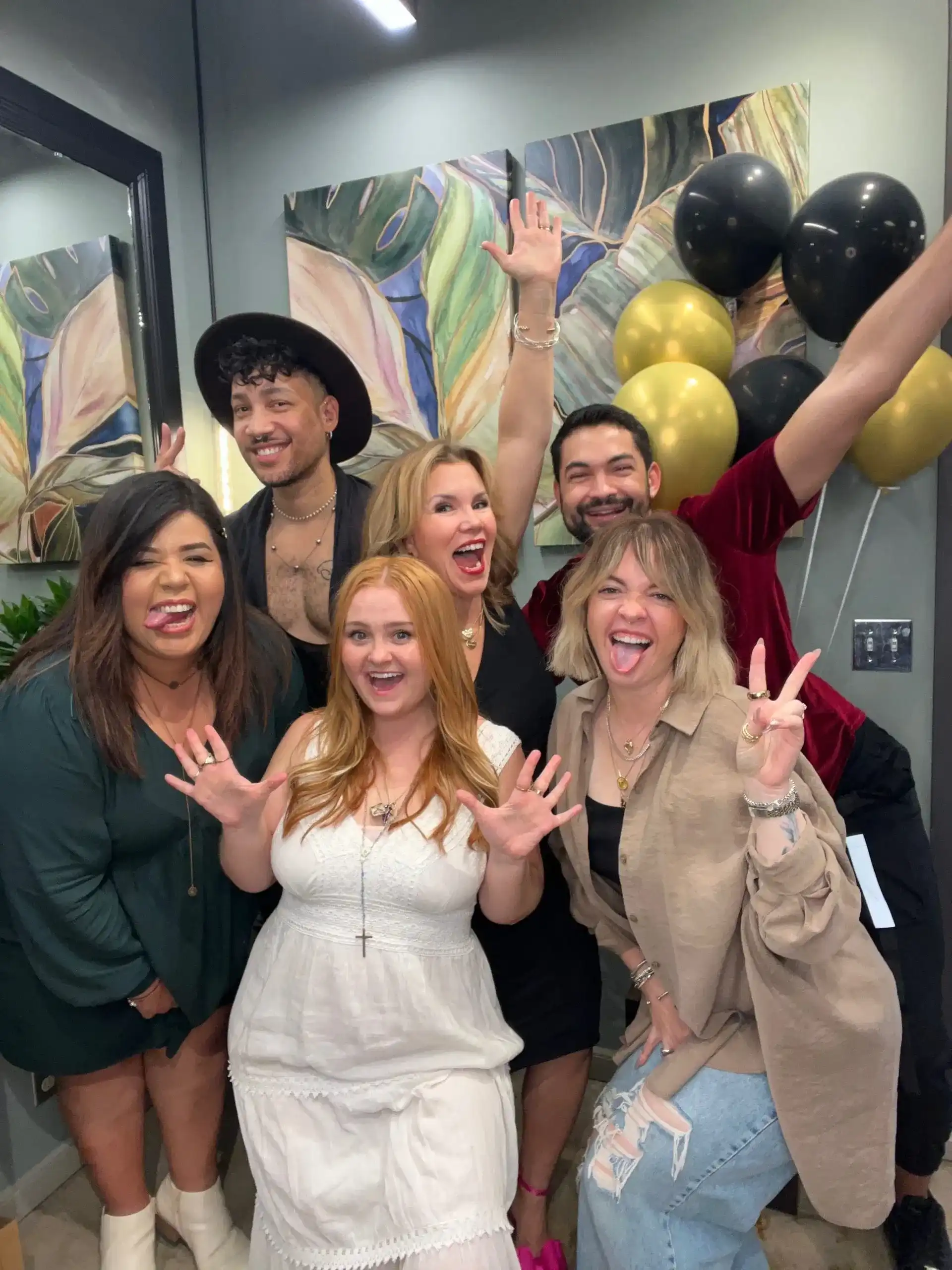 Group of six people smiling and posing playfully with balloons in the background.